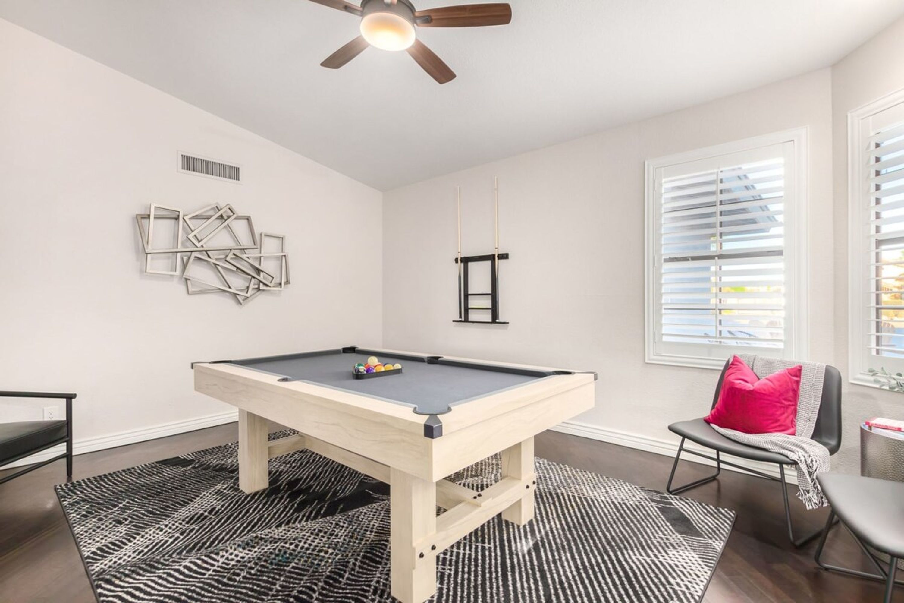 Une table de billard dans une pièce dotée d’un ventilateur de plafond, d’une fenêtre avec des stores et d’une chaise agrémentée d’un coussin rouge.