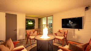 A cozy indoor seating area with a central fire feature, comfortable chairs, a flat-screen TV, and a visible room name sign. - Palmhouse Pool BBQ 5 Mins to Old Town (Scottsdale)