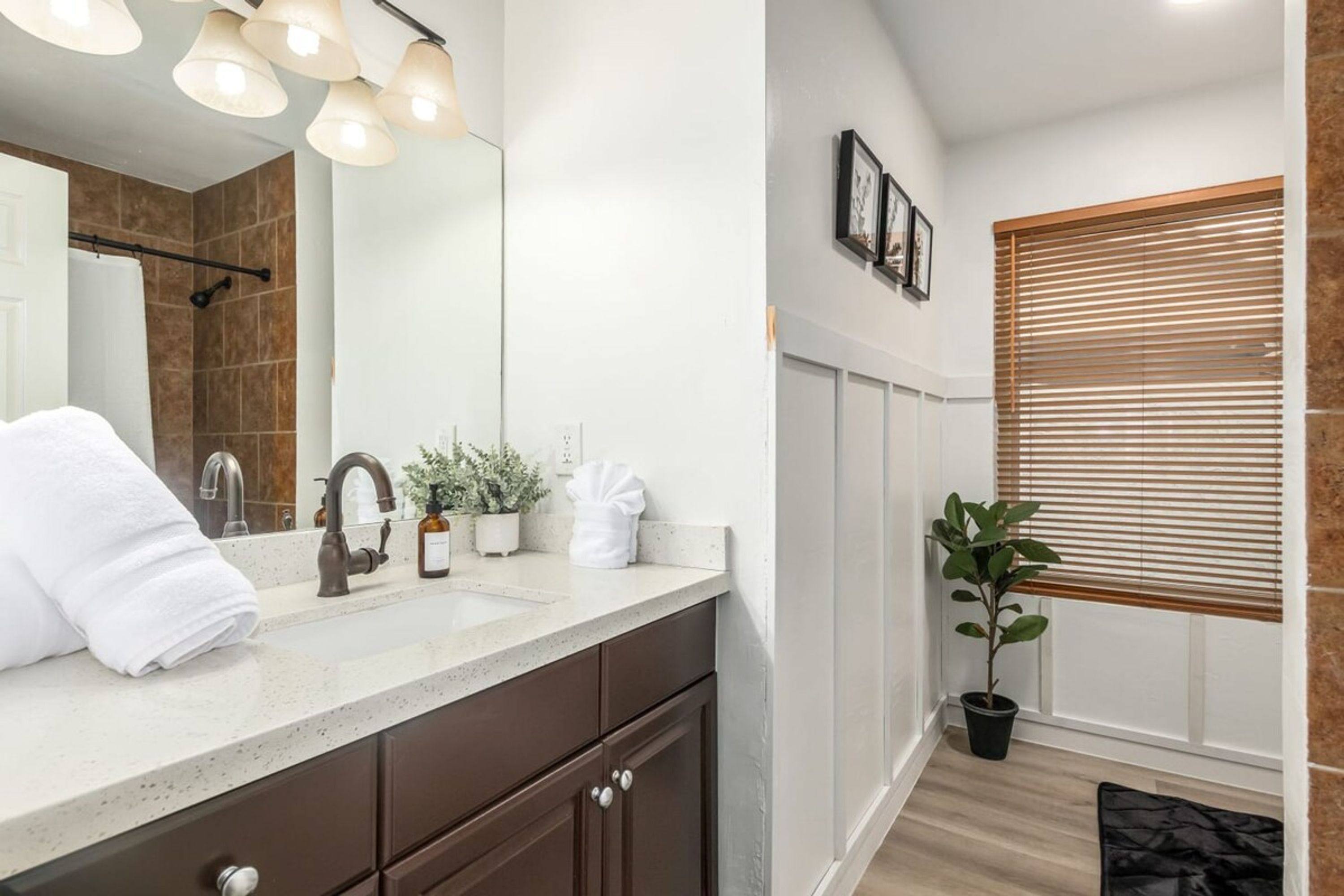 A modern bathroom with a large mirror, a sink, and a shower area. There are towels and toiletries on the counter, and a plant next to the window.