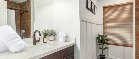 A modern bathroom with a large mirror, a sink, and a shower area. There are towels and toiletries on the counter, and a plant next to the window.