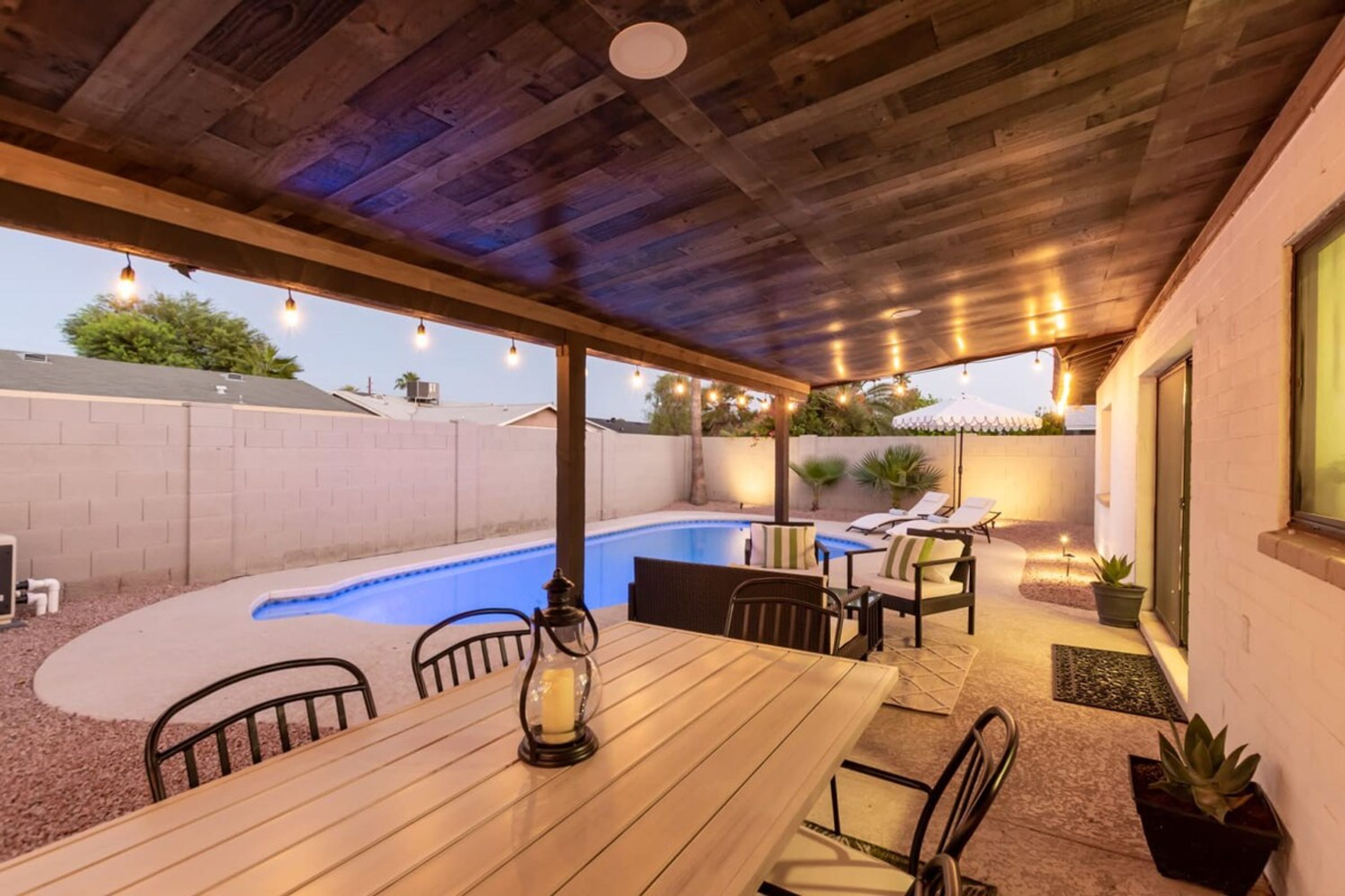 A covered patio area with a pool, wooden table, chairs, and potted plants.