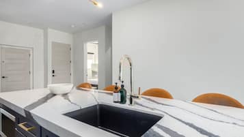 Una cocina moderna con encimera de mármol, un lavamanos negro y un grifo dorado. Se observan dos puertas al fondo y una silla con cojín.