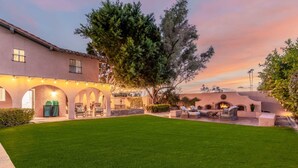 Un amplio patio al aire libre con un césped cuidado, mobiliario exterior y un gran árbol que proporciona sombra.