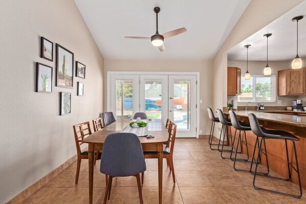 Une cuisine moderne avec un coin repas, comprenant un grand îlot central avec des tabourets de bar, un ventilateur de plafond et des tableaux encadrés accrochés au mur.