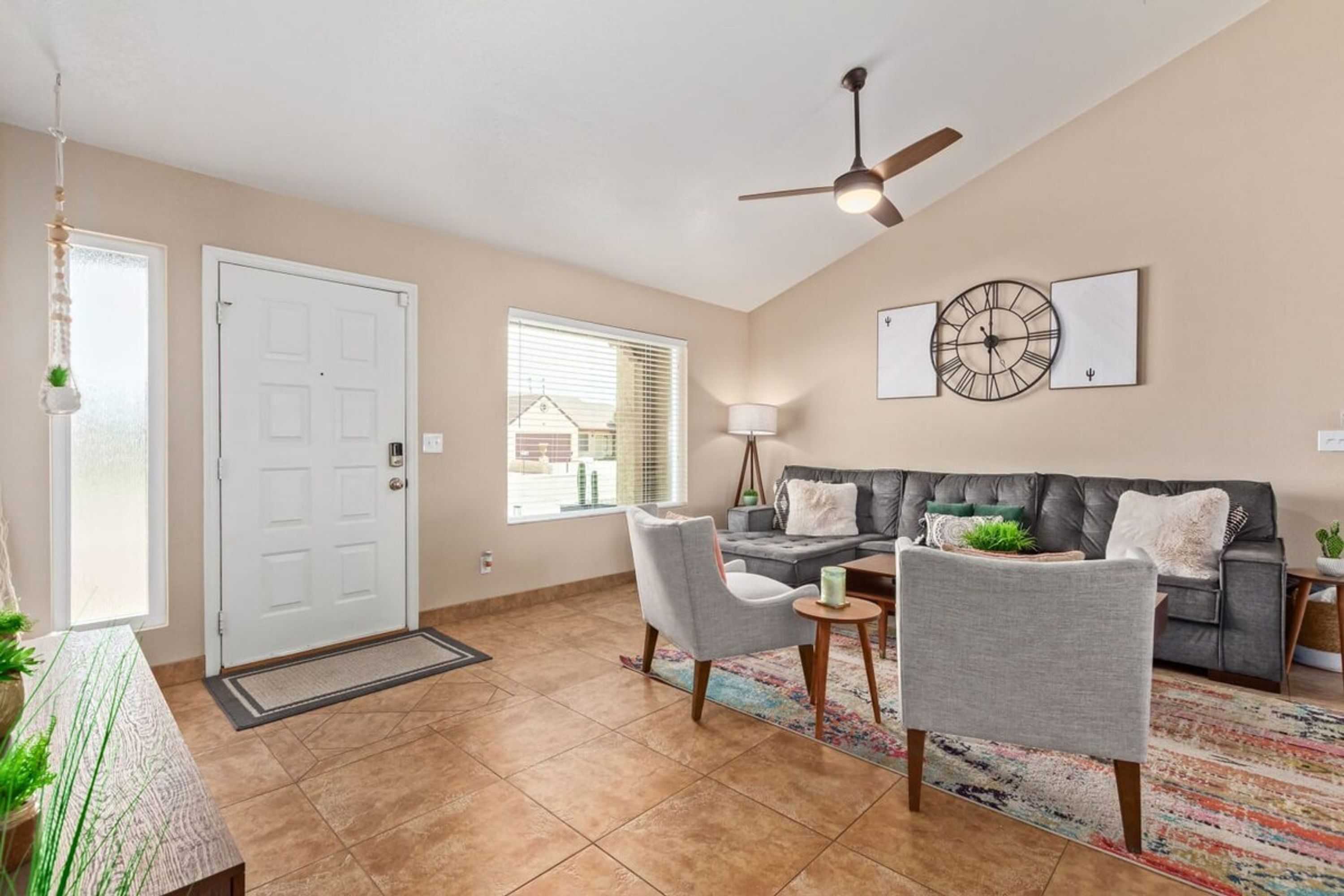A spacious living room with a grey sofa, two armchairs, a coffee table, and a ceiling fan. A large window provides natural light, and a door leads to the outside.