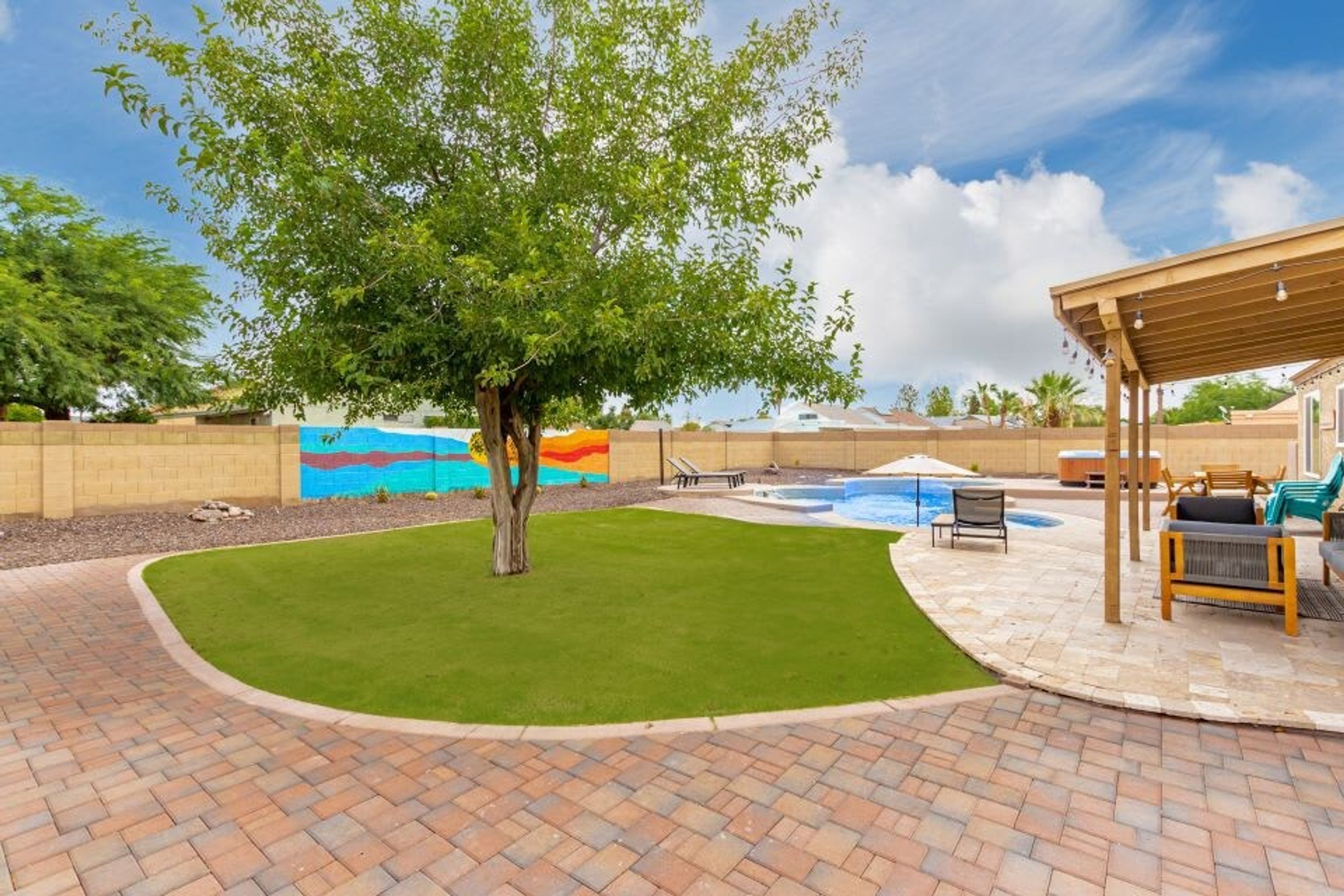 A backyard with a swimming pool, a covered seating area, and a mural on the wall.