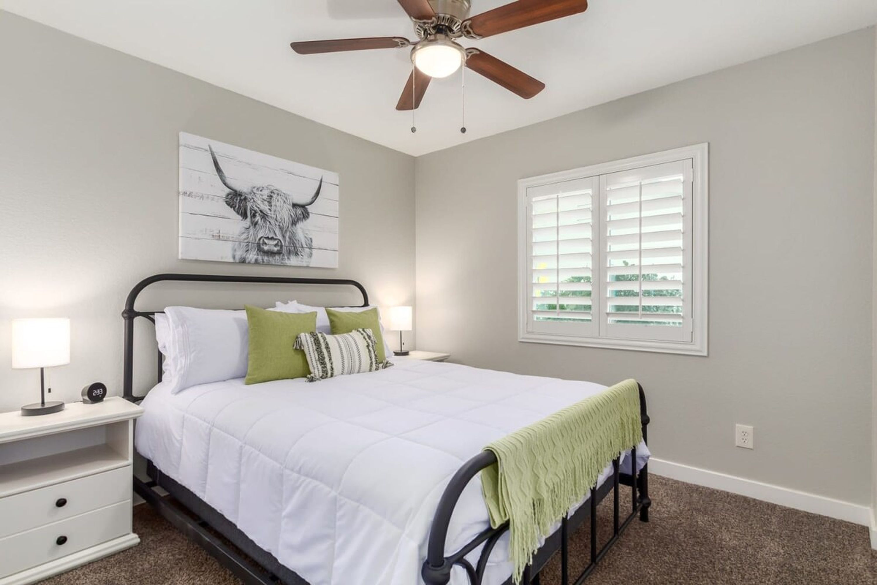 A bedroom with a bed, bedside tables, a ceiling fan, a window with blinds, and a wall-mounted artwork of a bull.