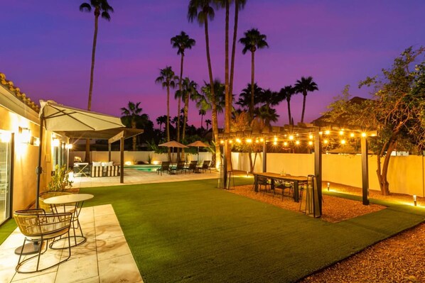 Un patio exterior bien iluminado con piscina, sillones de descanso y una pérgola con luces colgantes.