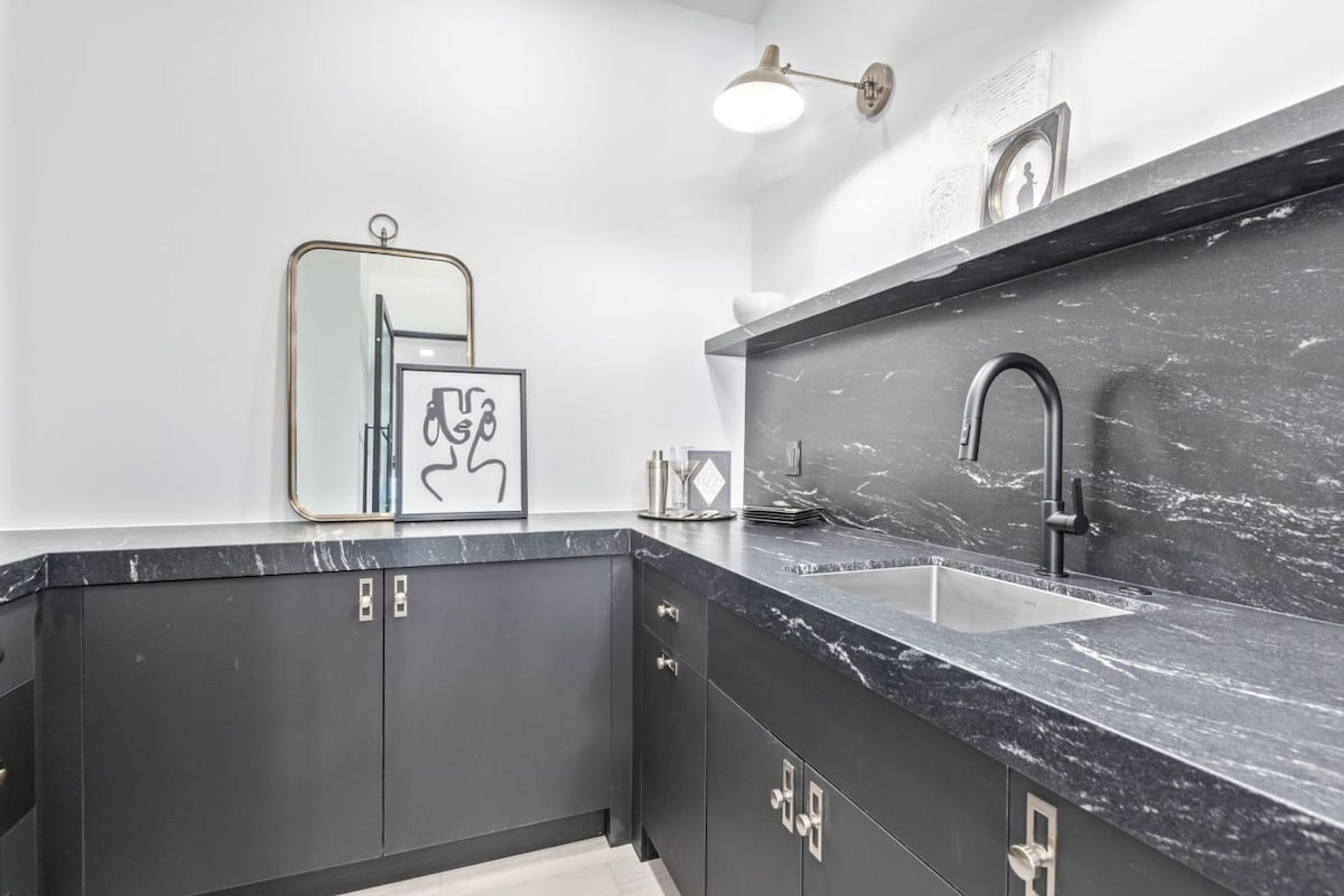 A modern kitchen with dark cabinets, a marble countertop, a sink, and a wall-mounted light fixture.