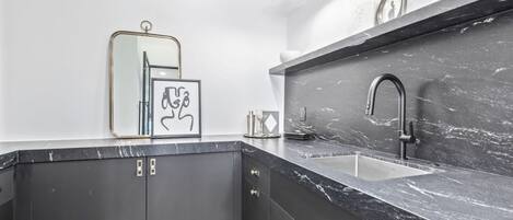 A modern kitchen with dark cabinets, a marble countertop, a sink, and a wall-mounted light fixture.