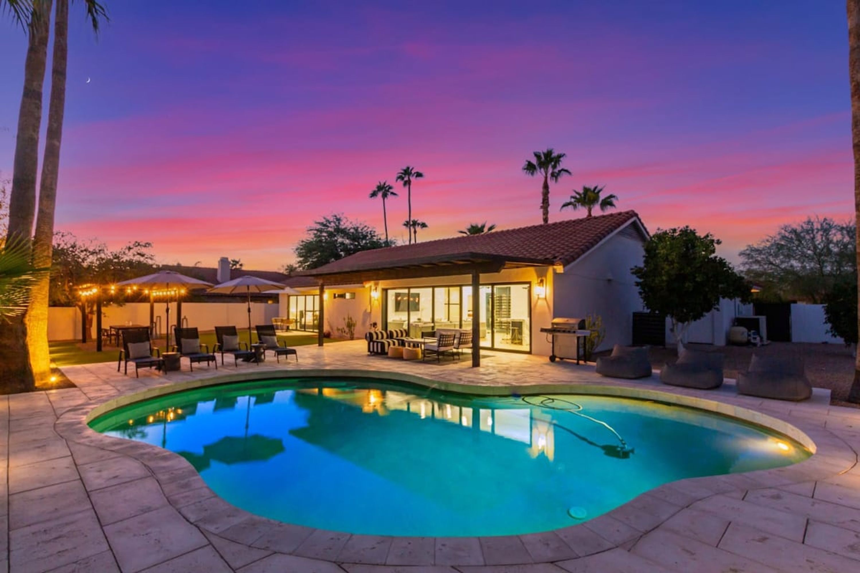 A swimming pool with a covered patio area, palm trees, and a sunset sky.