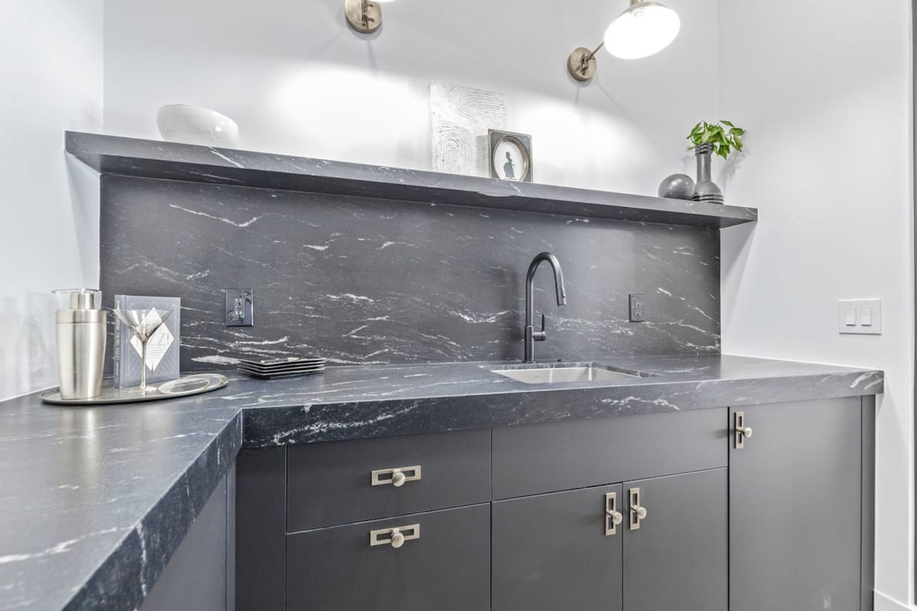A modern kitchen with a dark marble countertop, a sink, and a stainless steel faucet. There are cabinets with silver handles and a small potted plant on the countertop.