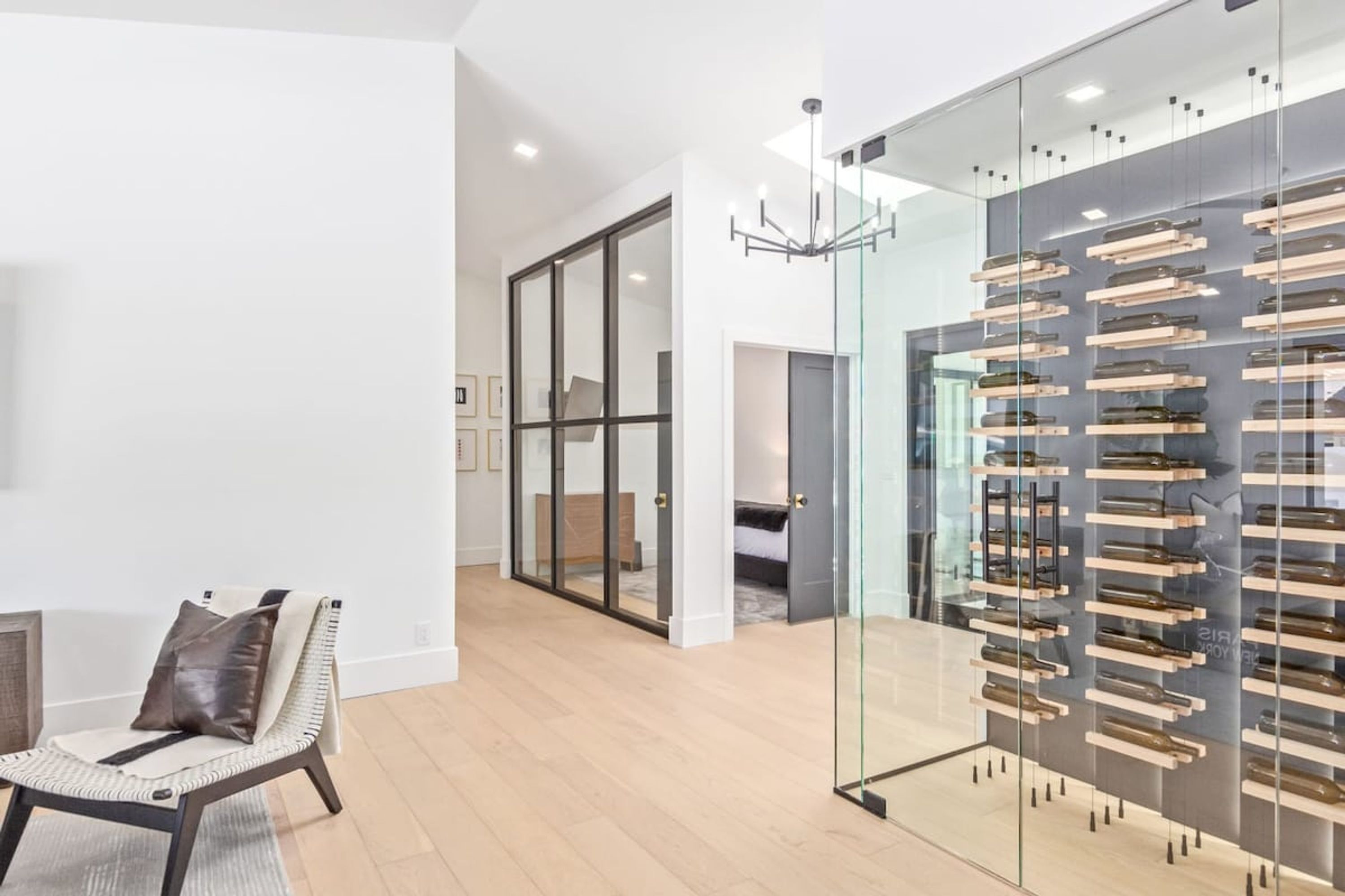 A modern interior with a glass partition displaying wine bottles, a seating area with a chair, and a hallway leading to another room.