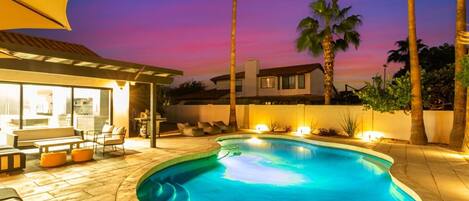 Une piscine à bord incurvé, entourée d’un patio aménagé avec des meubles d’extérieur et un espace salon abrité.