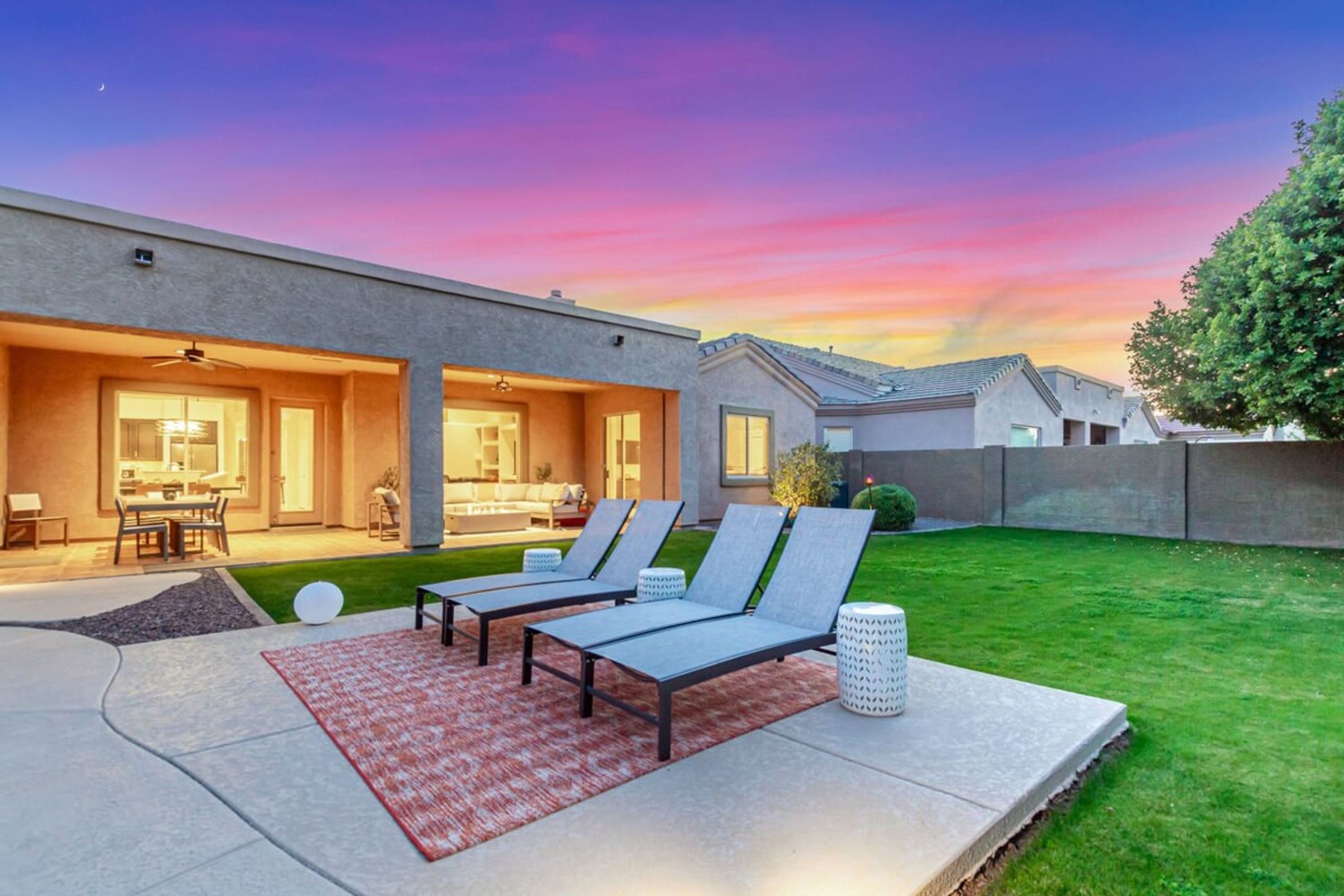 A modern house with a patio area featuring lounge chairs, a dining table, and a kitchen visible through the glass doors.