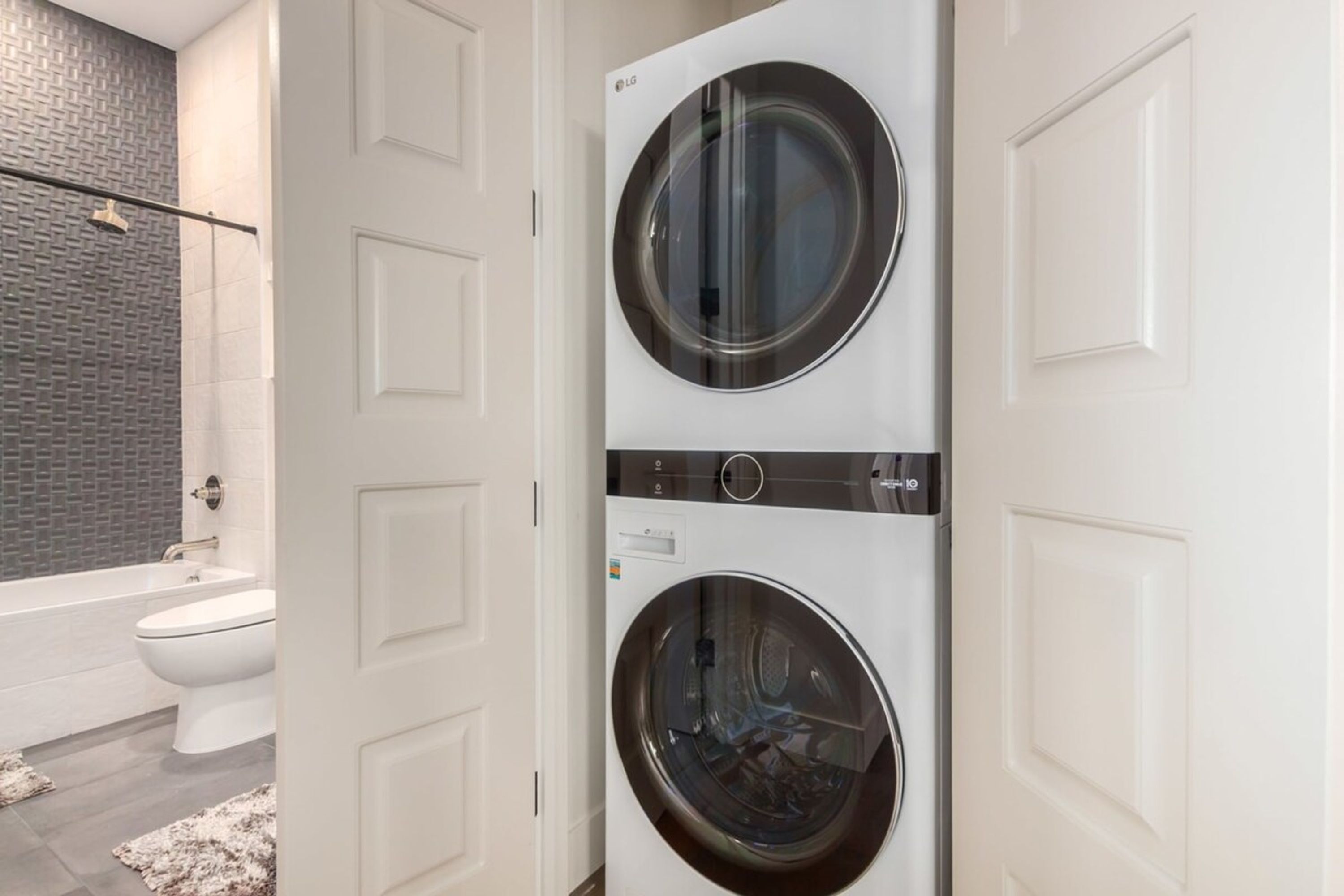 A laundry room with two front-loading washing machines, a white door, and a bathroom with a bathtub and toilet visible through an open door.