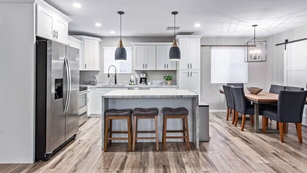 Une cuisine moderne avec des meubles blancs, un réfrigérateur en acier inoxydable, des tabourets en bois, des luminaires suspendus et un coin repas avec une table et des chaises en bois.