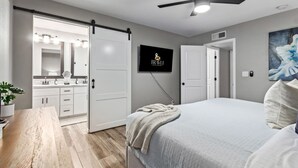 Une chambre moderne équipée d’un lit, d’un ventilateur de plafond, d’une télévision et d’une porte coulissante donnant accès à une salle de bain.