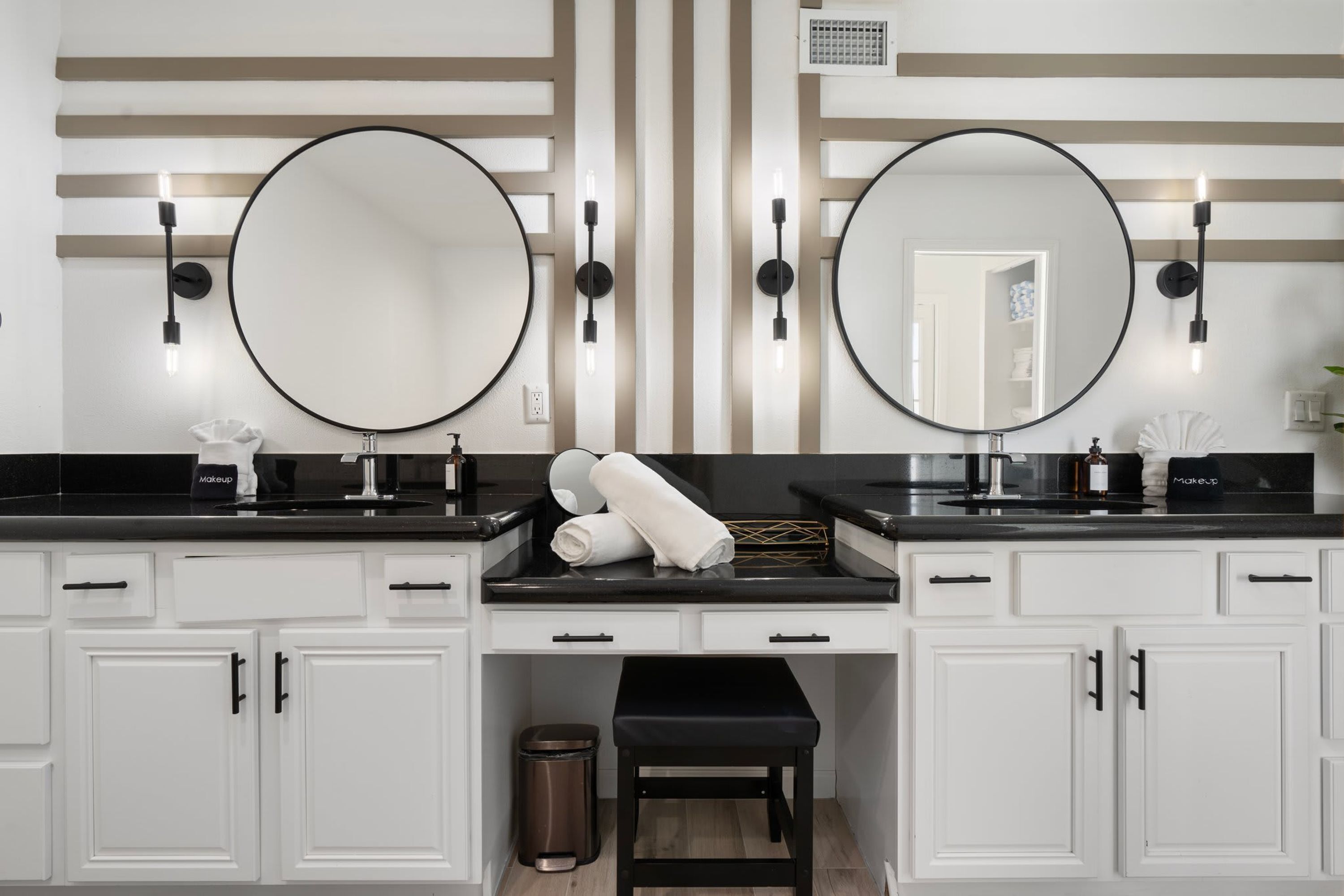 Une salle de bain avec deux lavabos, chacun doté d’un grand miroir et d’un luminaire au-dessus. On y trouve des meubles blancs sous les lavabos et un banc noir au centre.