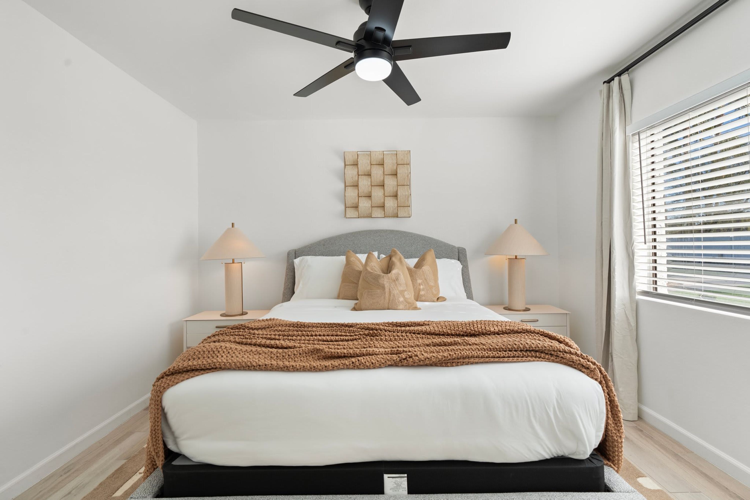 A modern bedroom with a bed, two bedside tables with lamps, a ceiling fan, a wall-mounted artwork, and a window with blinds.