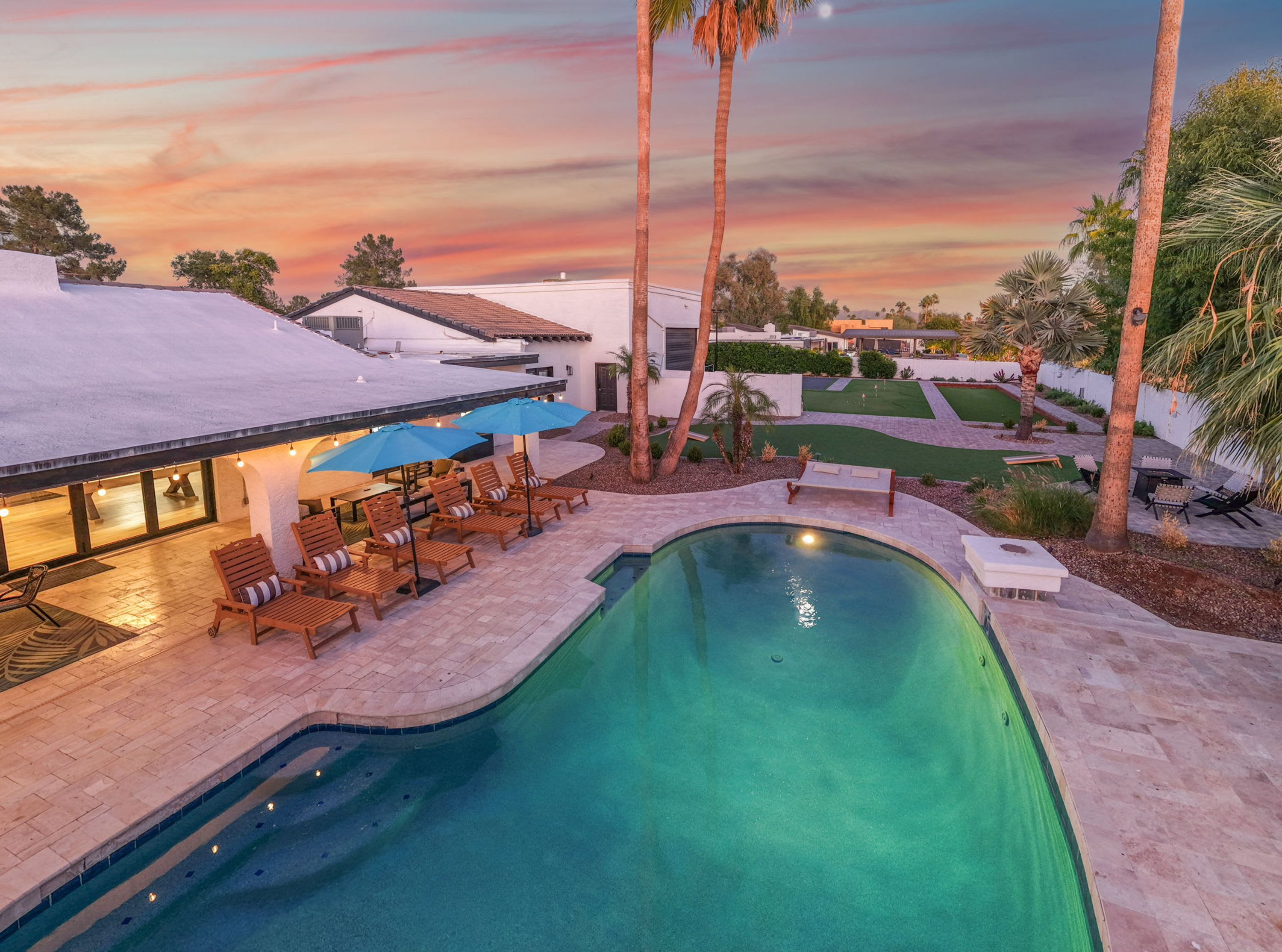 A swimming pool with lounge chairs and umbrellas, surrounded by palm trees and a well-maintained lawn.