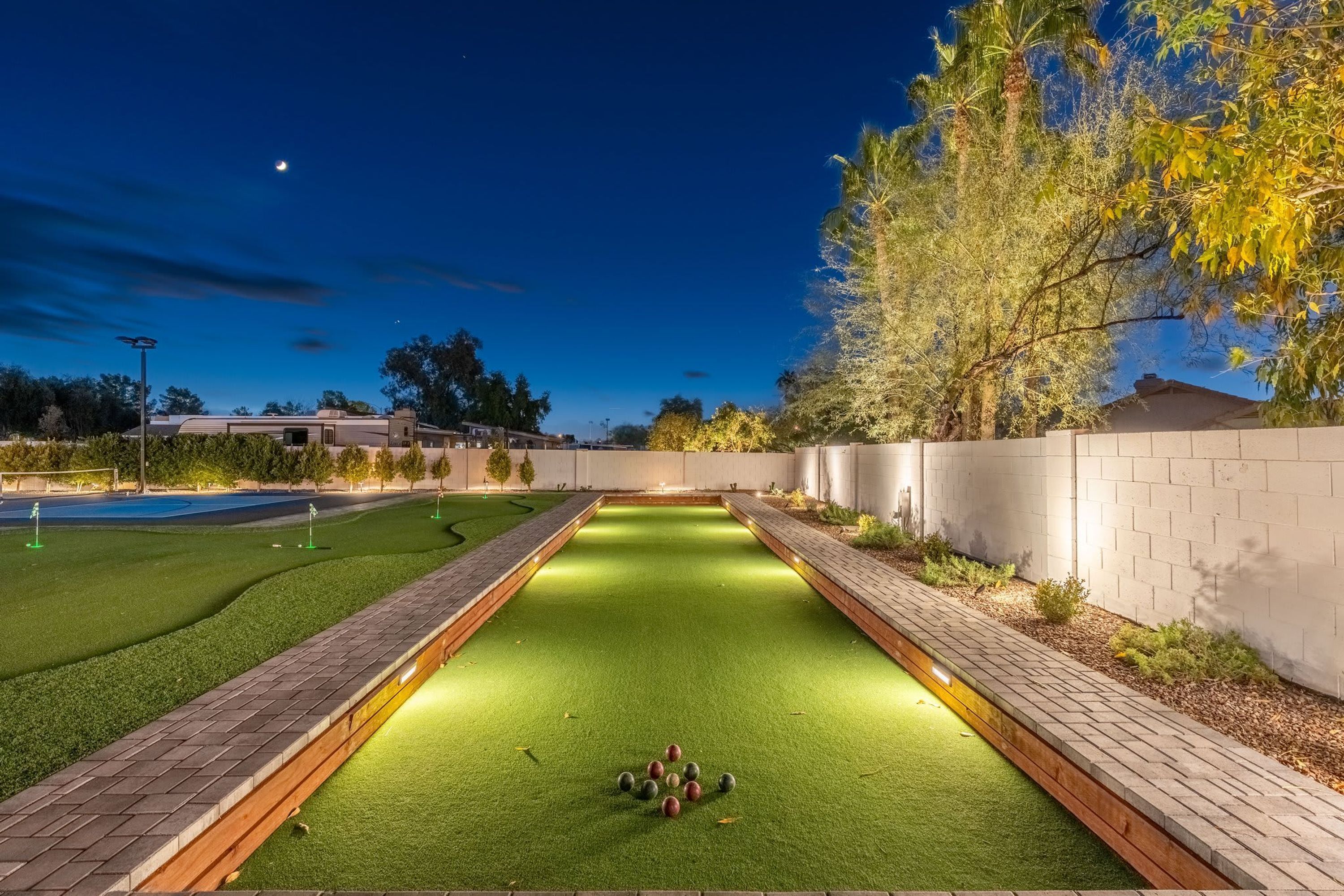 A well-lit croquet court at dusk with a clear view of the surrounding area, including a building and trees.