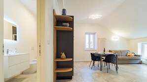 Un appartement moderne comprenant une cuisine, un coin repas et un salon. On y trouve une bibliothèque intégrée agrémentée d’objets décoratifs, une table à manger avec des chaises et un canapé.