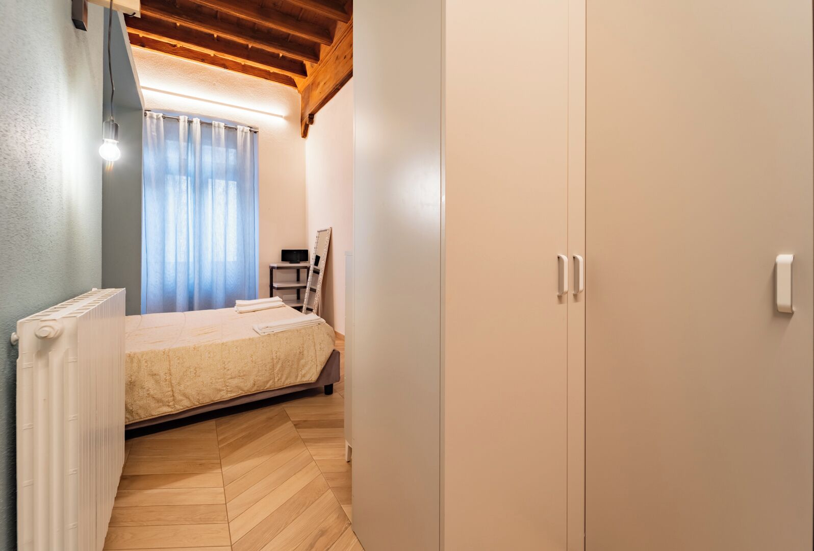 Ein Schlafzimmer mit einem Bett, einem Heizkörper, einer Holzbalkendecke, einem Kleiderschrank und einem Fenster mit Vorhängen.