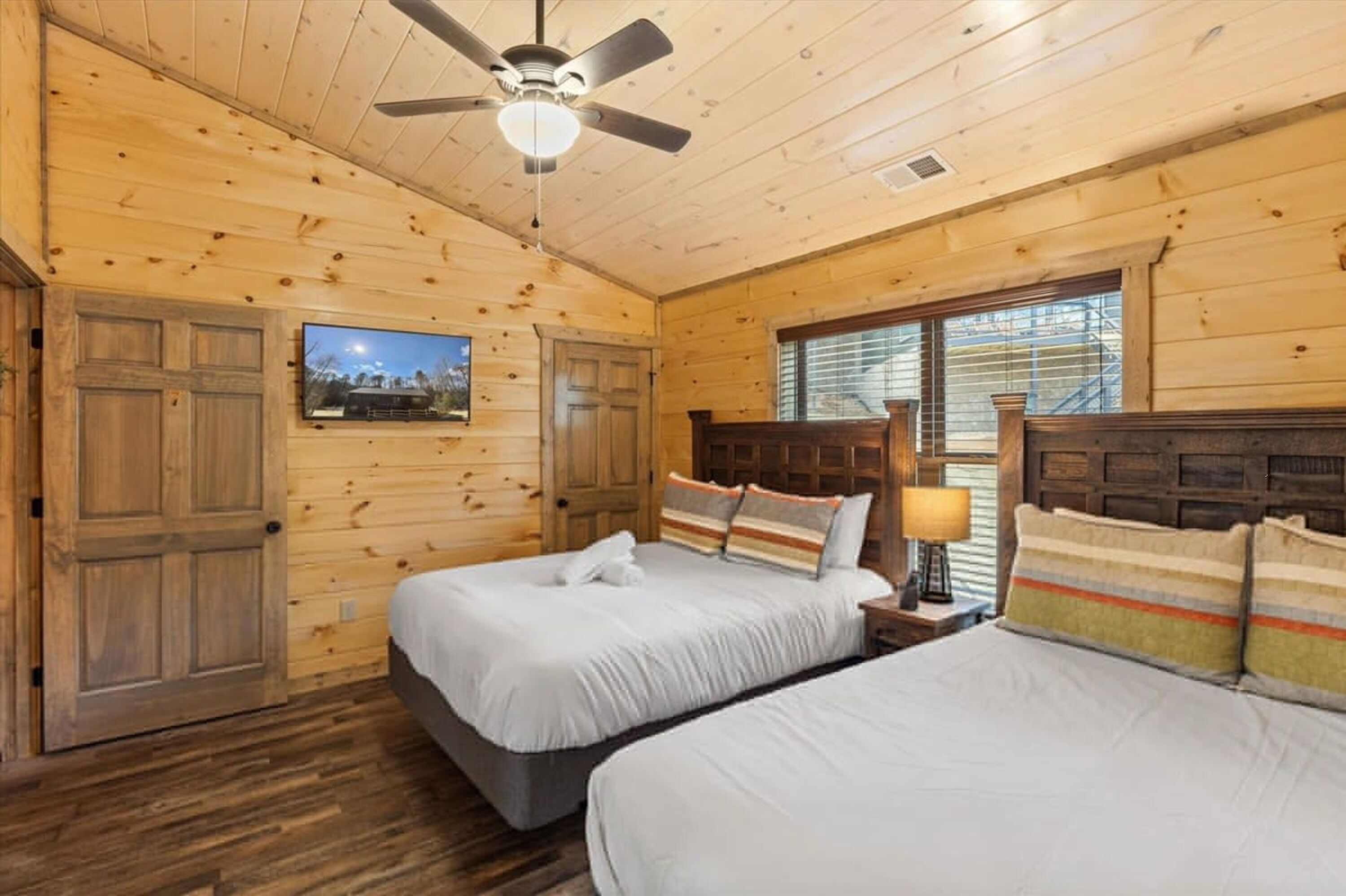 A wooden cabin bedroom with two beds, a ceiling fan, a TV, and a window with blinds.
