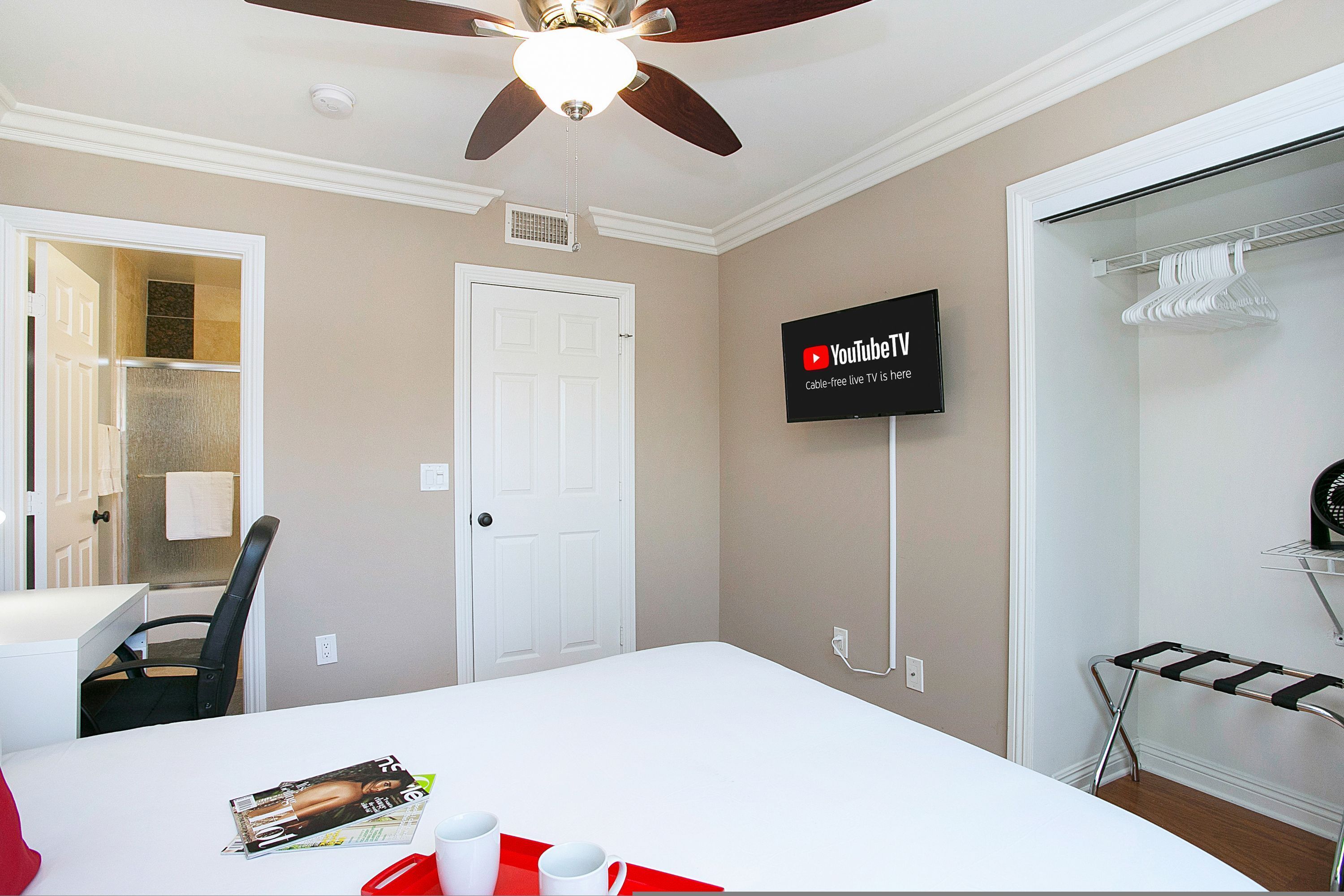 A hotel room with a bed, a TV mounted on the wall, a closet with a hanging rack, and a ceiling fan.