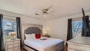 A bedroom with a large bed, a ceiling fan, a nightstand with a lamp, and a dresser.
