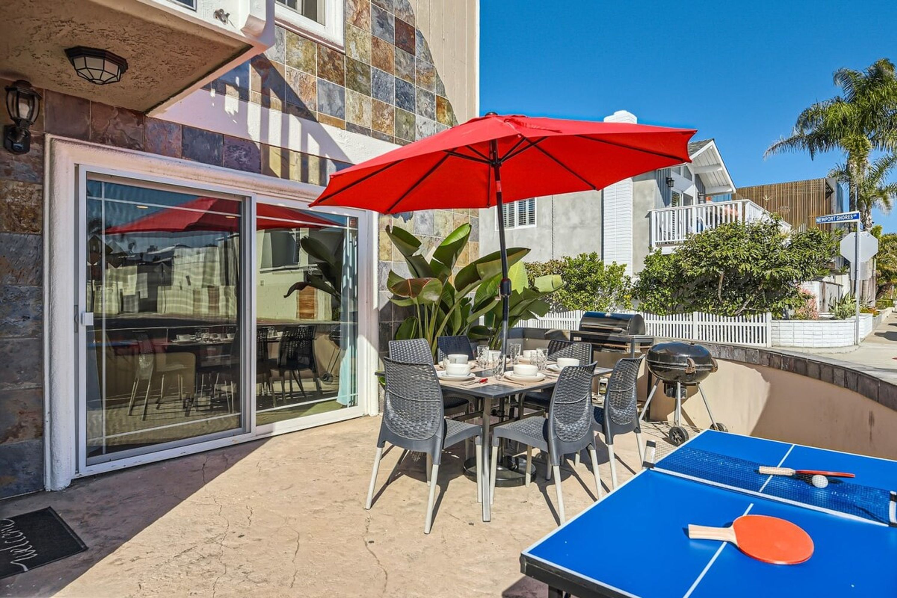 Un espace extérieur aménagé pour les repas, avec une table de ping-pong, des chaises et un parasol rouge.