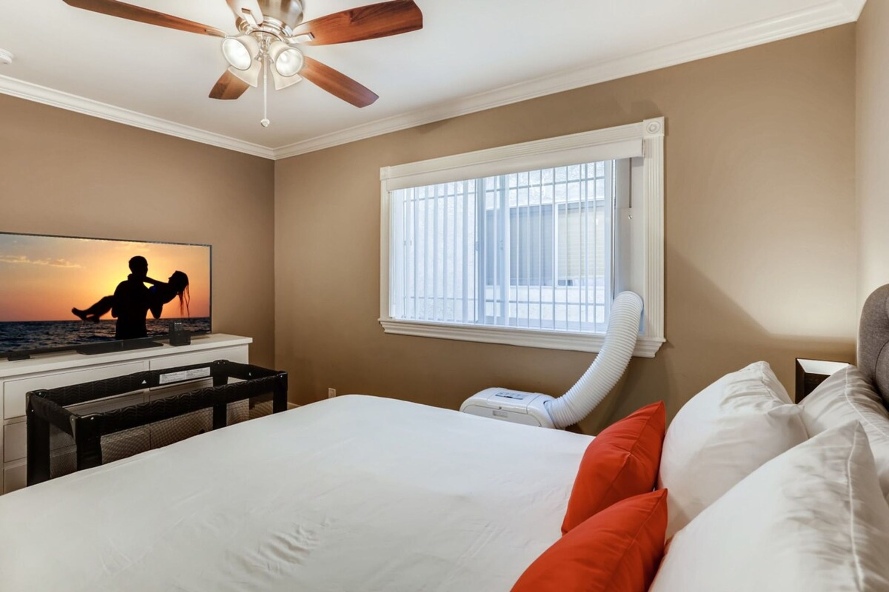 Un dormitorio con cama, almohadas, ventilador de techo, ventana con persianas y un televisor que muestra una escena de atardecer.