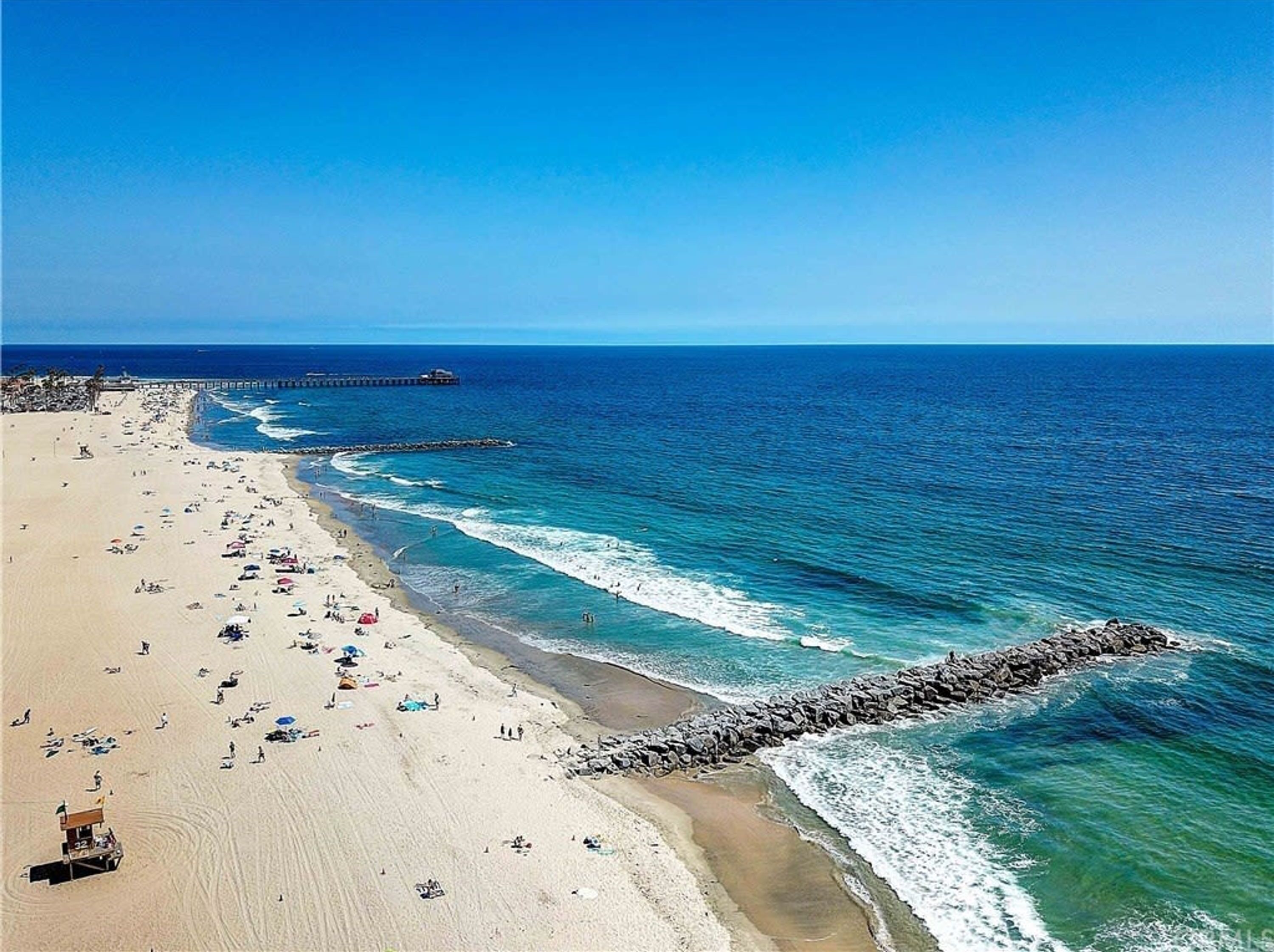 A beach with a rocky breakwater, clear blue water, and a pier in the distance.