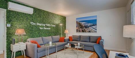 A modern living room with a green wall, grey sofas, a coffee table, a desk with a laptop, and a beach view painting.