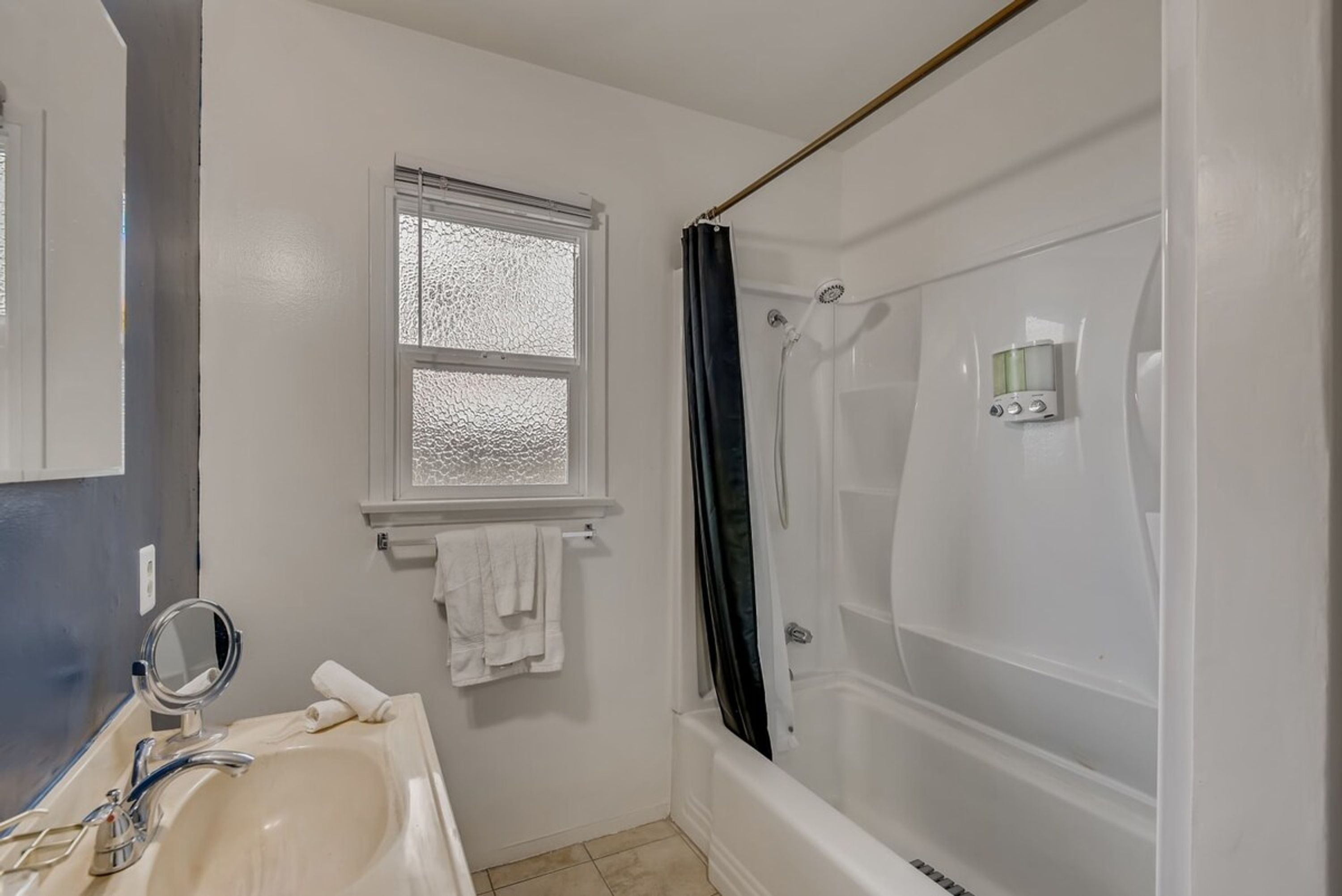 A bathroom with a white sink, a mirror, a window with frosted glass, a shower with a curtain, and a towel rack.