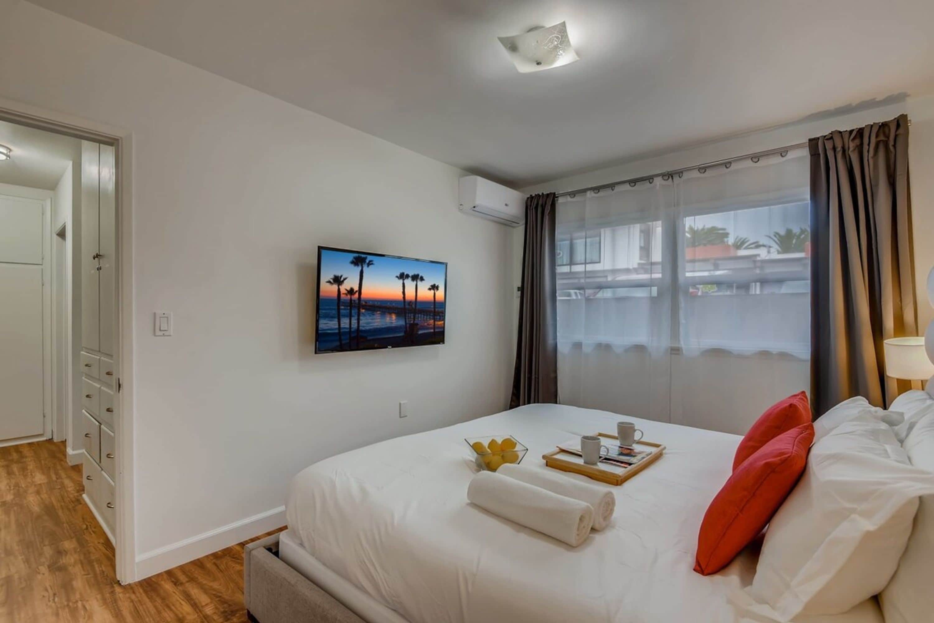 A hotel room with a bed, a painting of a sunset, a window with curtains, and a view of buildings outside.