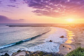 A coastal scene with a curved walkway along the beach, a pier extending into the water, and a sunset over the horizon. - Waking up in Your own 3BR Beach Home (Newport Beach)