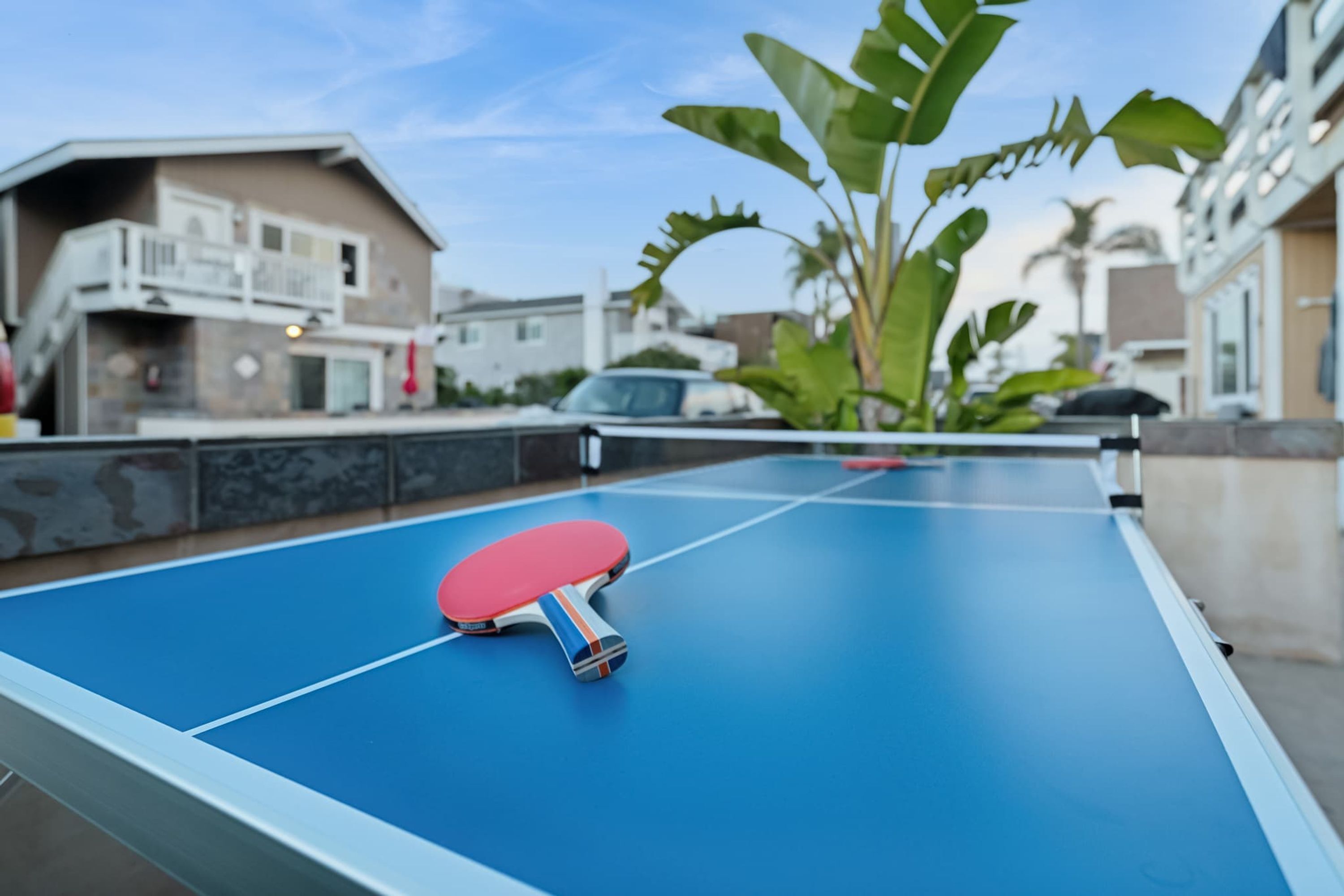 Ein Tischtennis-Tisch mit einem roten Schläger im Vordergrund, vor einem blauen Hintergrund. Im Hintergrund sind Wohnhäuser und ein geparktes Auto zu sehen.