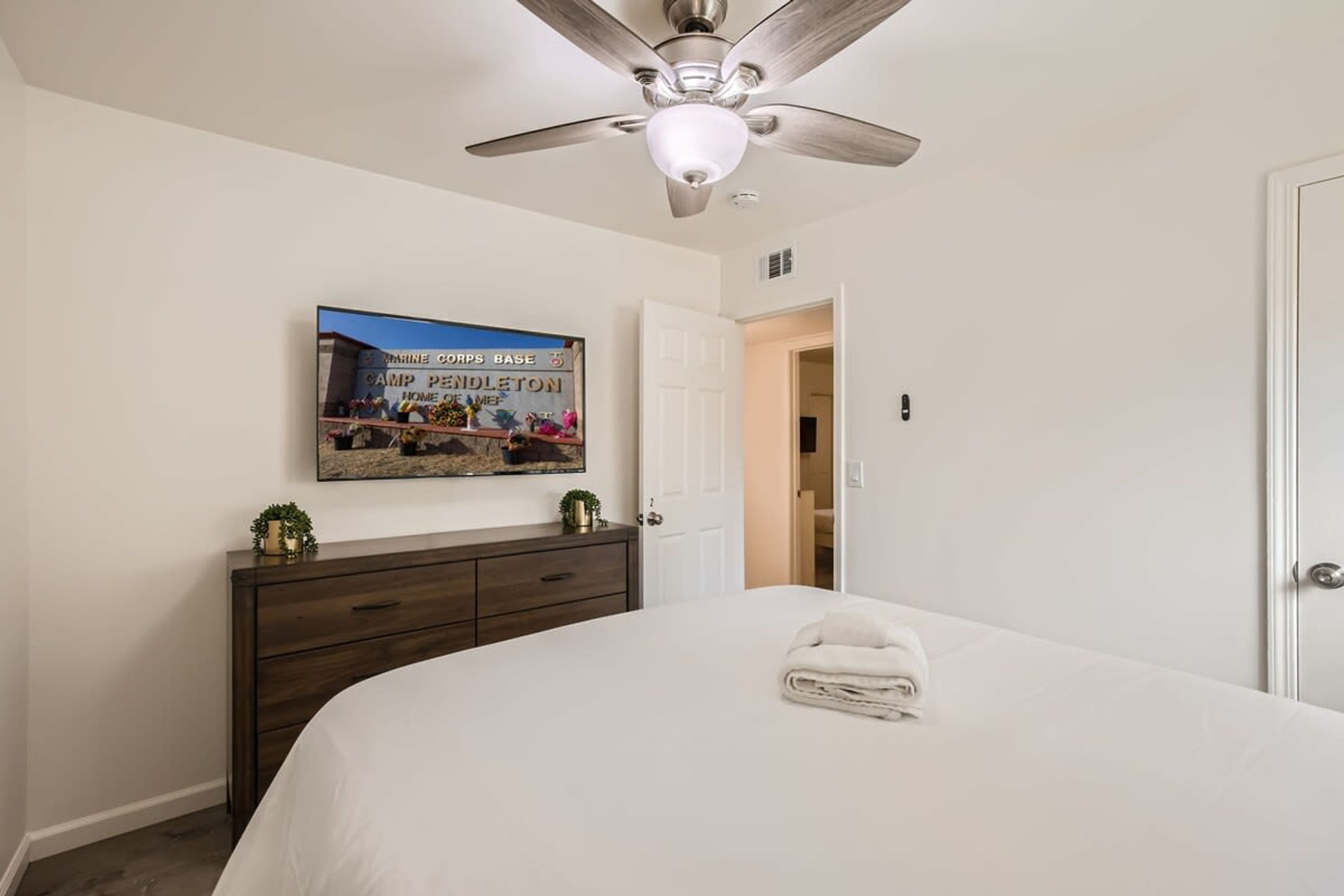 A bedroom with a bed, a ceiling fan, a dresser, a framed picture on the wall, and a door leading to another room.