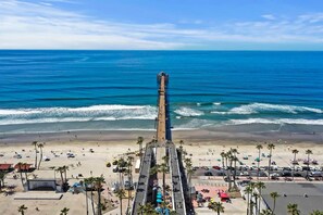 A coastal view with a pier extending into the ocean, a sandy beach with umbrellas, and palm trees. - Awesome 6BR - 15 Min To Beach (Oceanside)