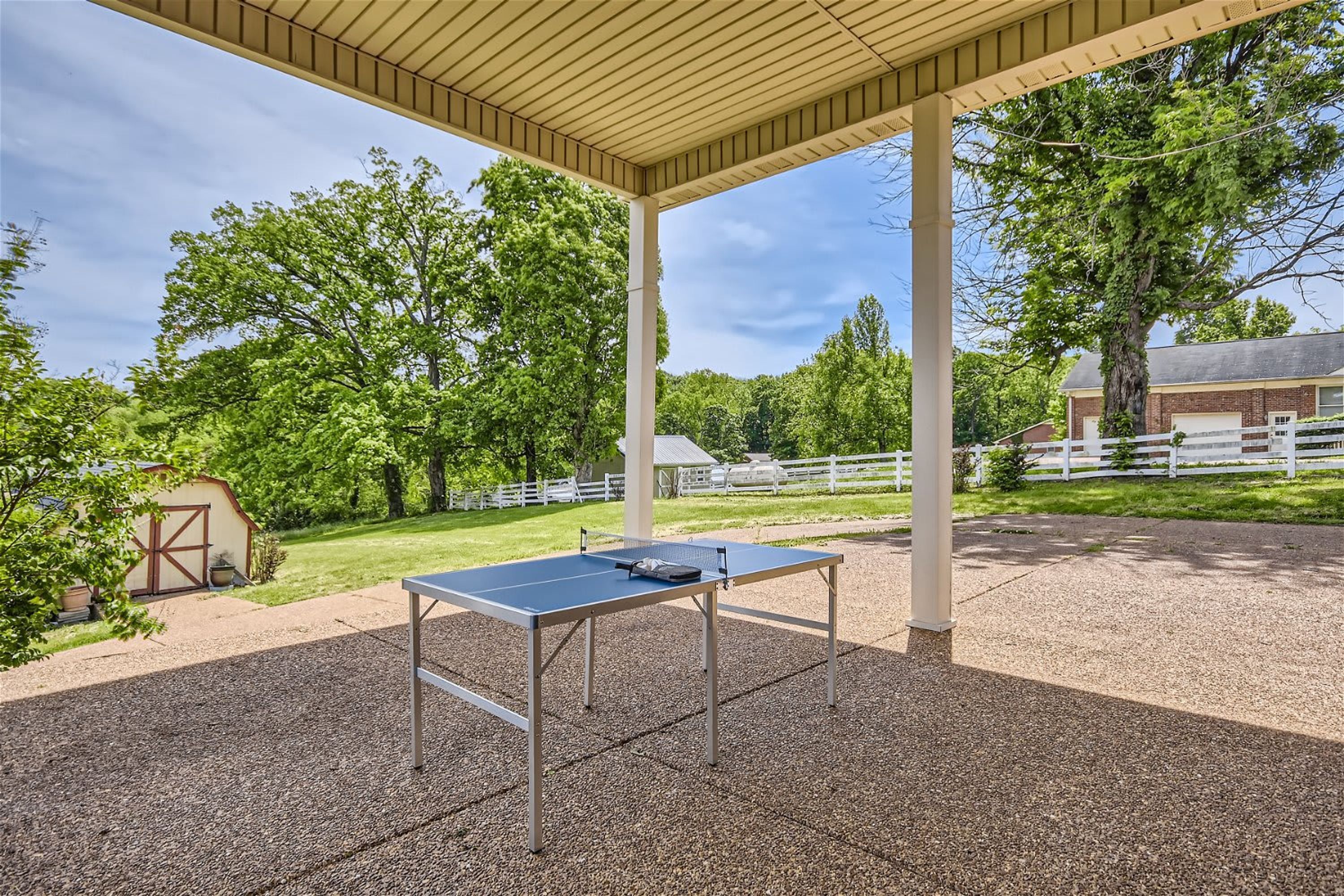 An outdoor ping pong table under a wooden pergola with a gravel surface, surrounded by trees and a white fence.