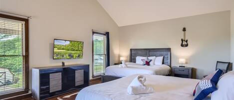 A modern hotel room with two beds, a flat-screen TV, a ceiling fan, and a view of greenery through the windows.