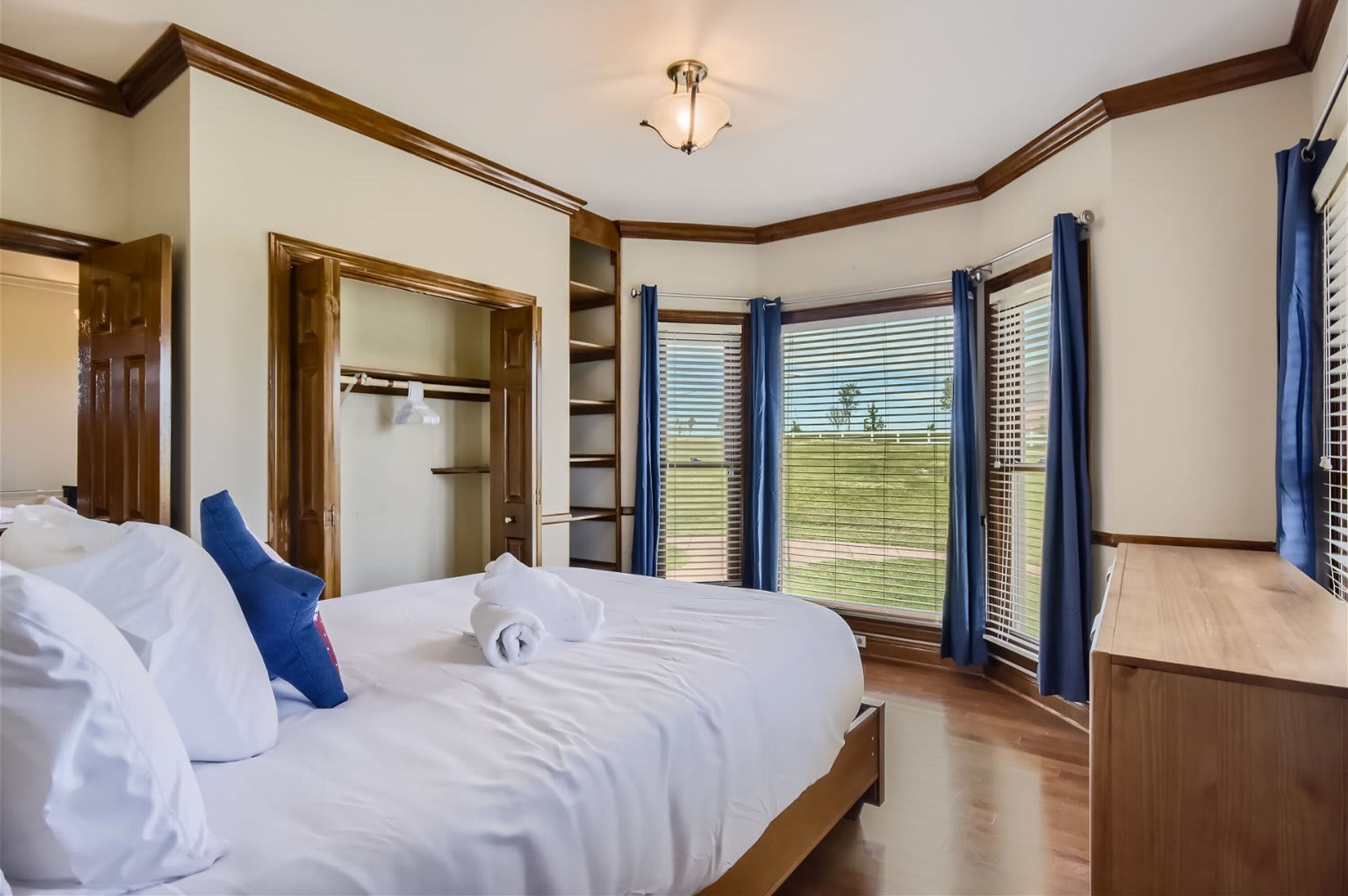 Un lit bien fait, avec du linge de lit blanc et un coussin bleu, une table de chevet en bois, un grand miroir et une vue sur un espace extérieur verdoyant, visible à travers une fenêtre munie de stores.