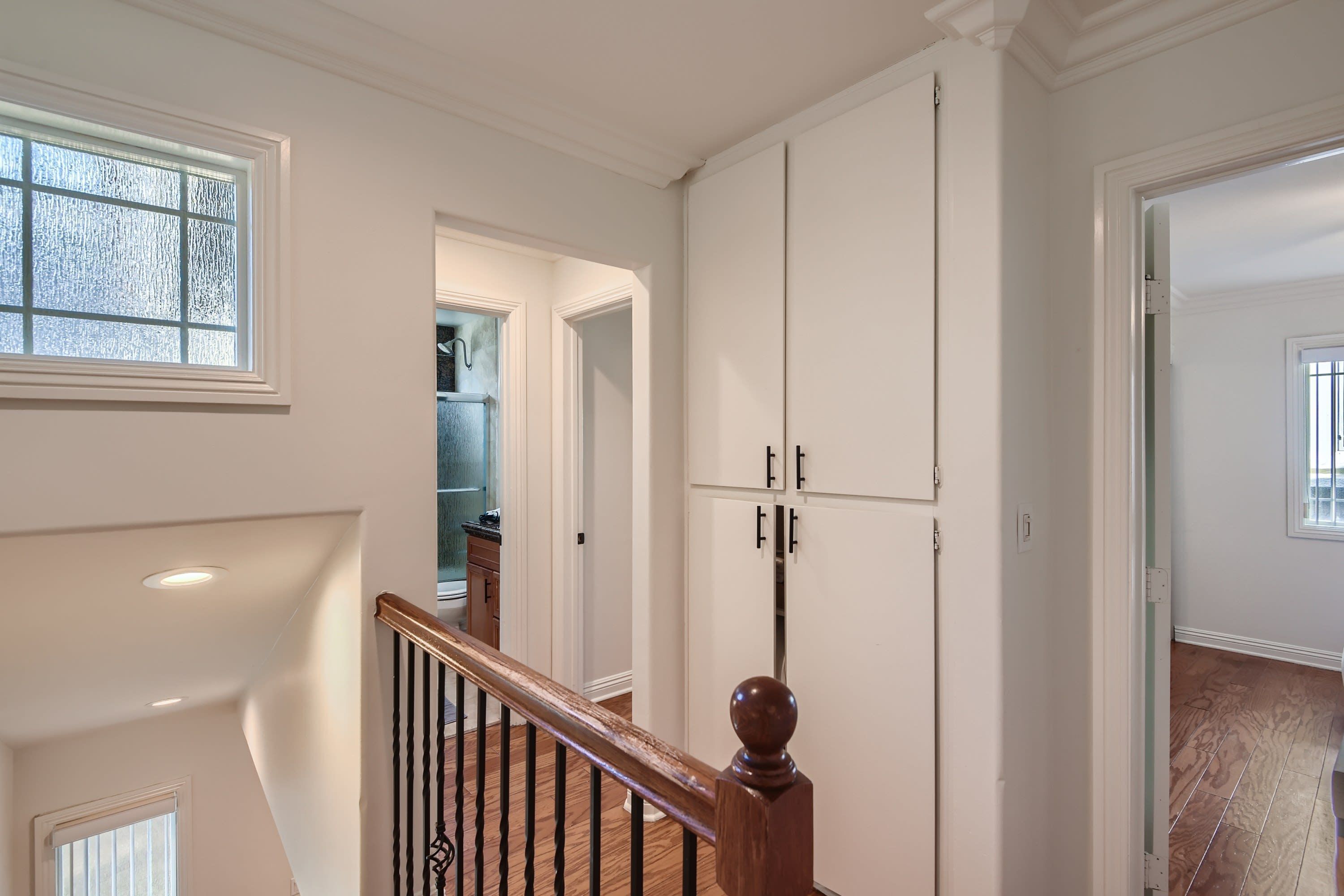 A staircase with a wooden handrail and black balusters, leading to a room with white walls and a large white cabinet.