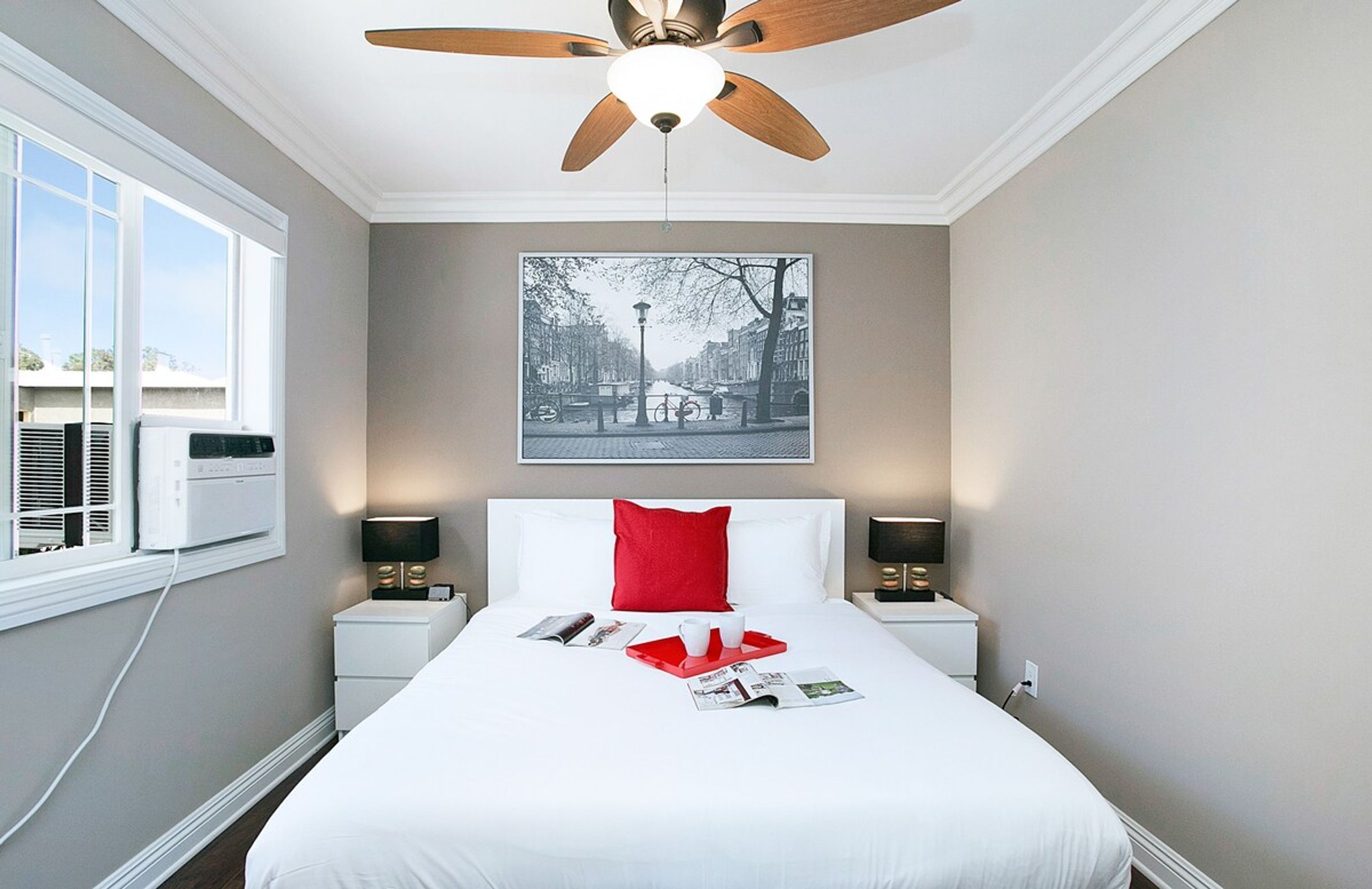 Quarto de hotel com cama, ventoinha no teto, janela com ar condicionado, mesa de cabeceira com abajur e quadro pendurado na parede.