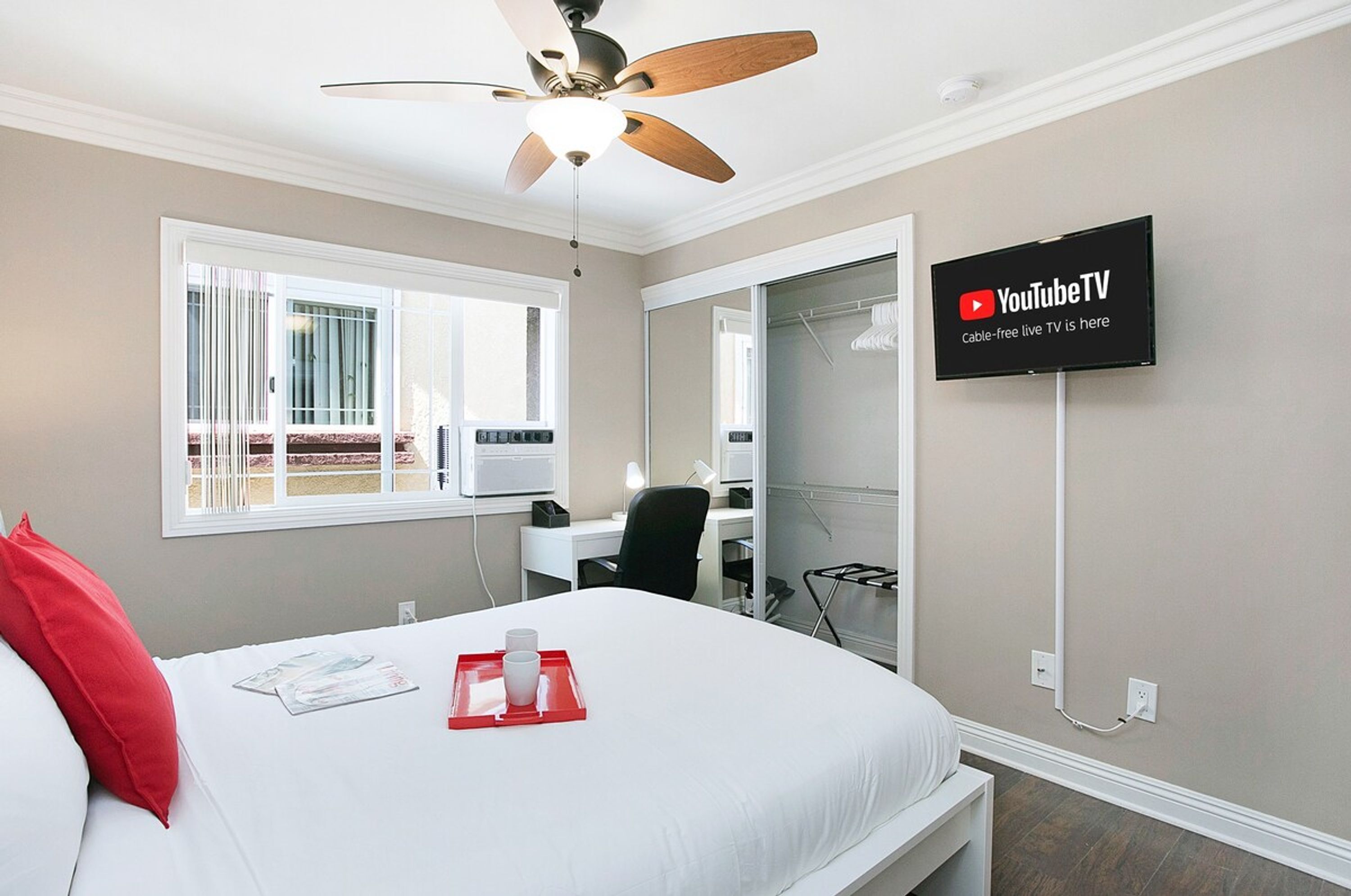 A hotel room with a bed, a ceiling fan, a desk with a chair, a TV mounted on the wall, and a window with blinds.