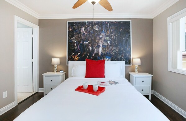 A hotel room with a large bed, two bedside tables with lamps, a red pillow, and a tray with two cups.
