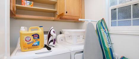 A laundry area with a washing machine, a dryer, a basket, and a shelf with folded clothes and a box of detergent.