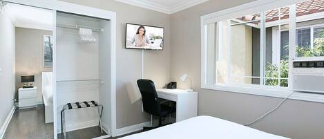 A hotel room with a bed, a desk, a chair, a TV, a window with an air conditioner, and a sliding door leading to a bathroom.